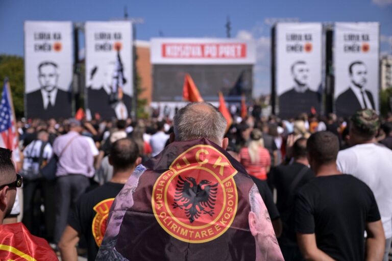 Демонстрант завиткан во знаме со амблемот на ОВК на собир во Косово, пред големи портрети на поранешни лидери на организацијата.